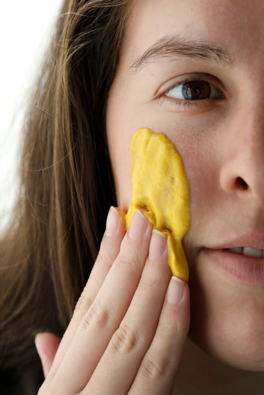 Woman applying a bright yellow turmeric face mask paste to the face by hand.