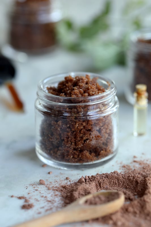 A vanilla body scrub using brown sugar, cacao powder, sunflower oil, and vanilla extract is stored in a clear glass salve jar and used for exfoliating and nourishing the skin.
