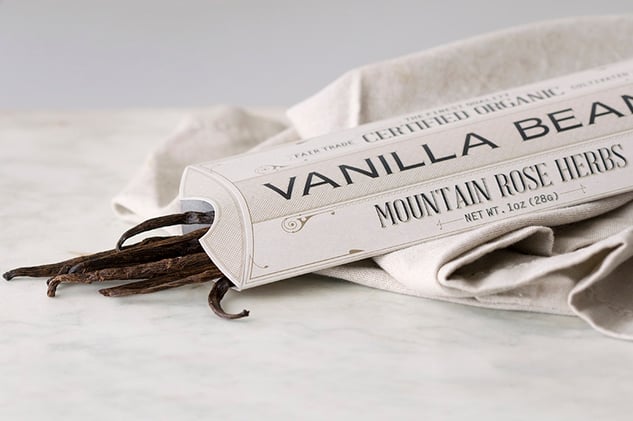 Rectangular paper package holding a bundle of vanilla beans lying on counter with fabric
