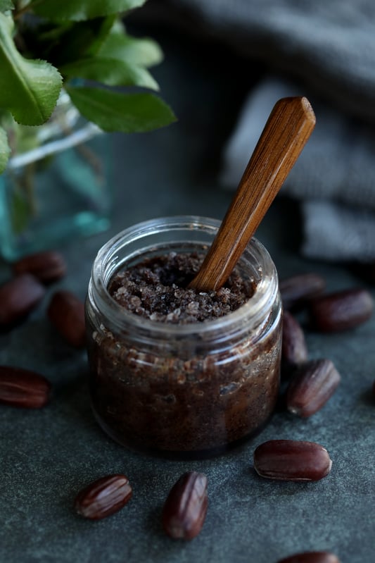 A DIY body scrub with vanilla and jojoba meal, honey, and coarse sea salt in a small glass salve jar. A wooden spoon is sitting in the salt scrub. Fresh jojoba beans are scattered around. 
