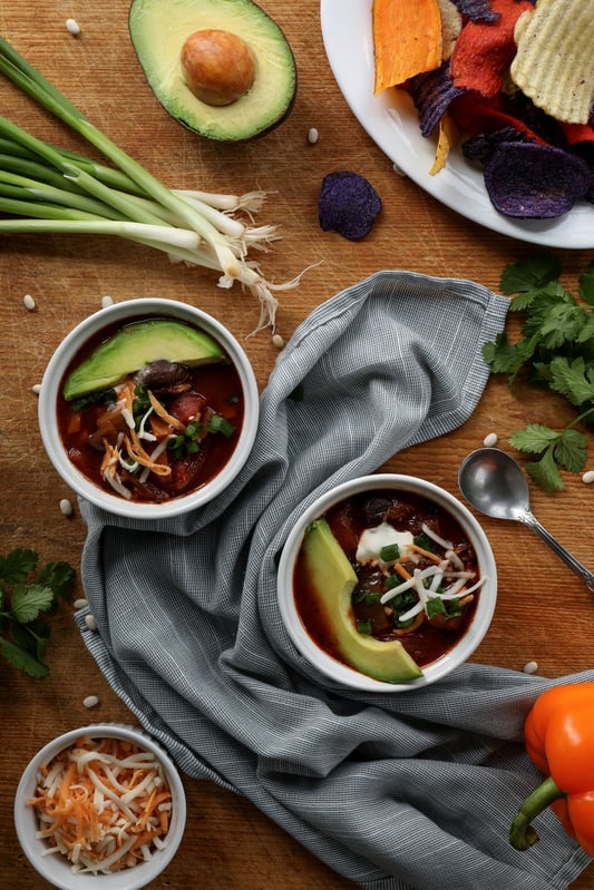 Bowls of hearty vegan chili are ready to be served, topped with vegan cheese, vegan sour cream, and organic avocado slices. Other ingredients include fresh green onion, bell peppers, cilantro, and baked vegetable chips as a side dish.