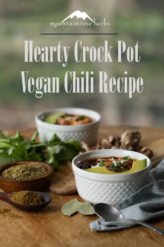 Bowls of hearty vegan chili are ready to be served, topped with vegan cheese, vegan sour cream, and organic avocado slices. Other ingredients include fresh green onion, bell peppers, cilantro, and baked vegetable chips as a side dish.