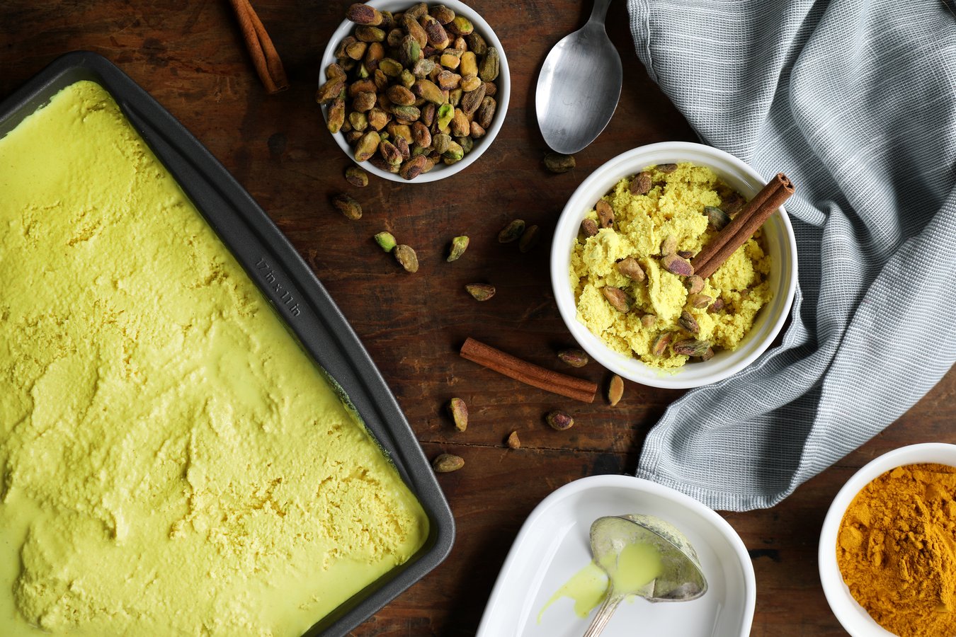 Ingredients to make vegan golden milk ice cream, including golden milk turmeric powder, cinnamon sticks, pistachios with a pan of finished ice cream and a scoop.
