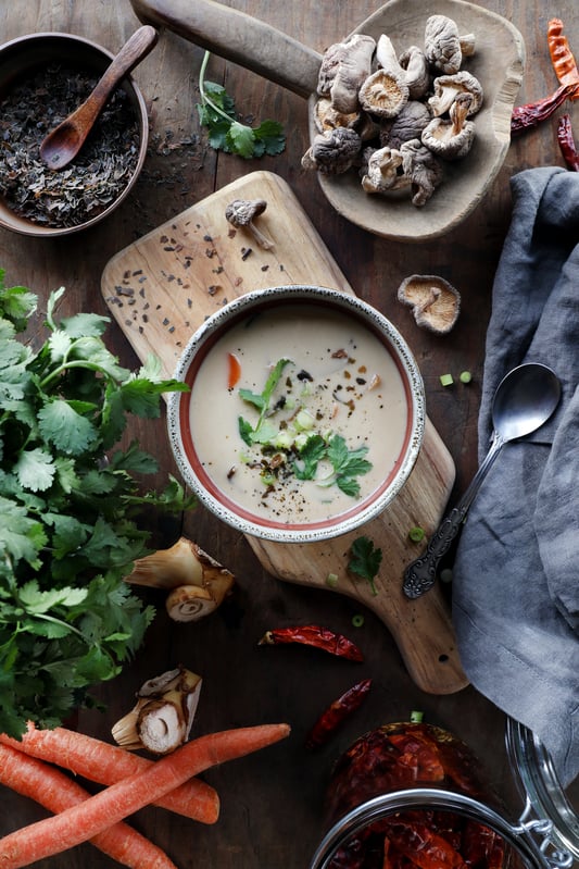 Cup of creamy tom kha thai coconut soup surrounded by fresh ingredients and herbs. 