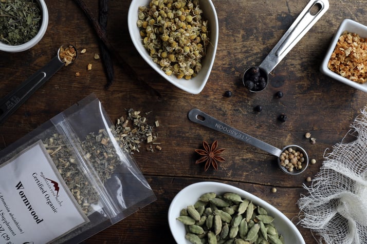 Ingredients for making homemade vermouth laying in bowls on counter including cardamom and wormwood and orange peel