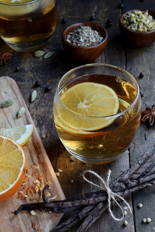 Light beverage with lemon slice in it displayed with vanilla beans, citrus and other spices. 