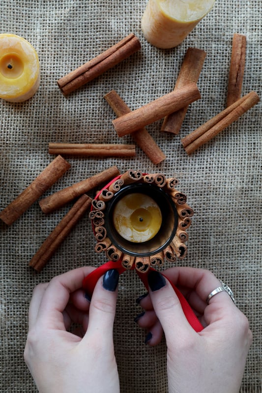 Hand tying red bow around cinnamon sticks bunched around candles.  Displayed cinnamon sticks and yellow candles around hands on burlap backdrop.