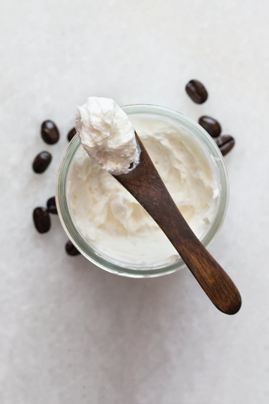 Overhead view of wooden spoon with whipped body butter on glass jar of butter surrounded by coffee beans