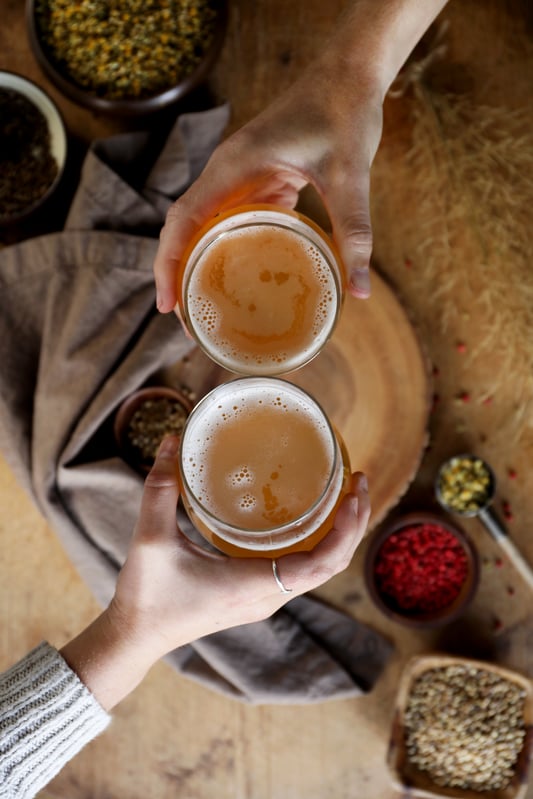 Two people are clinking glasses of frothy golden witbier among various herbal brewing ingredients like chamomile, coriander, and pink peppercorns. 