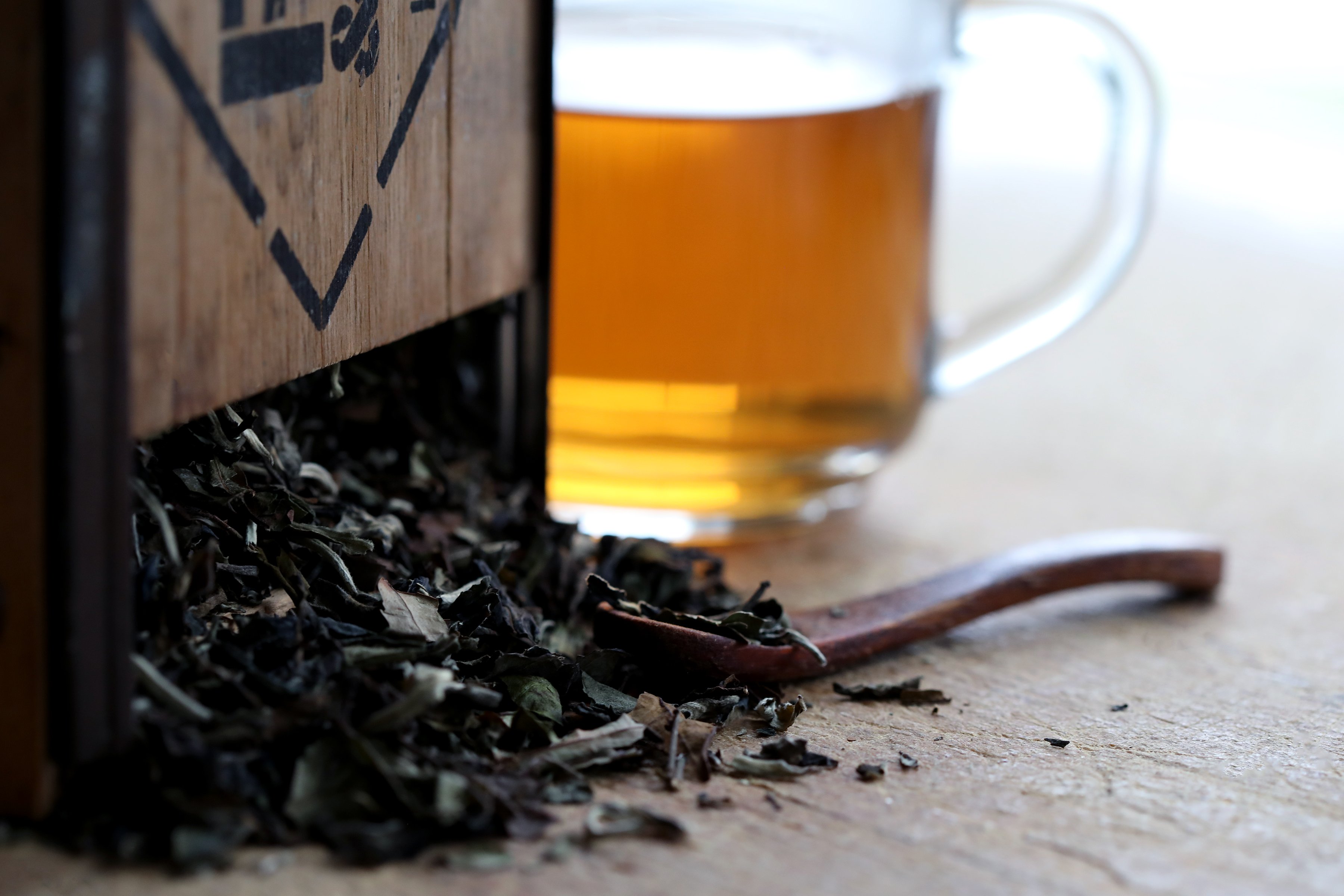 Dried white tea leaves pouring out of a rustic wooden box with spoon. Mug of brewed tea in background, golden in color.