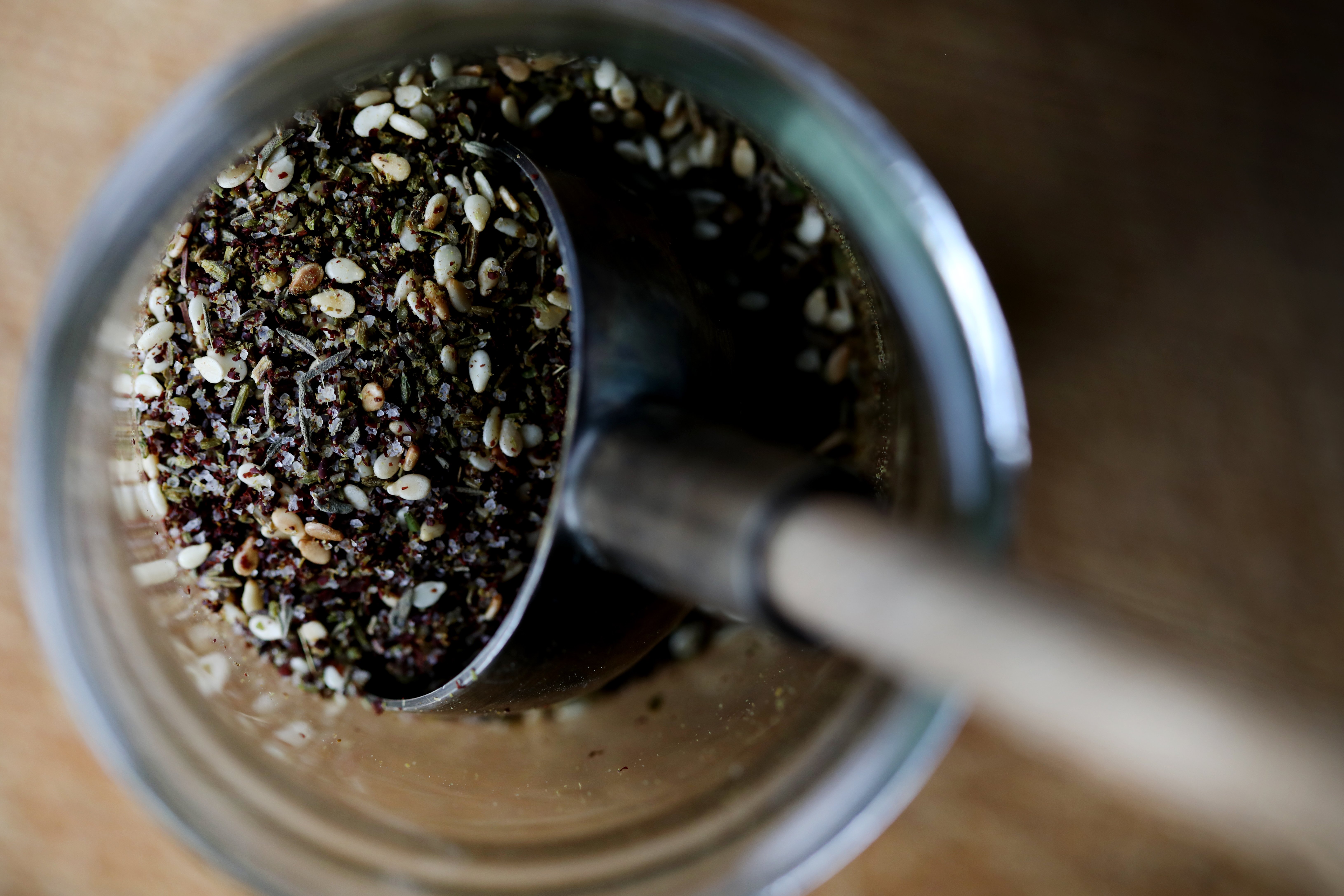 Detail of a metal scoop lifting an aromatic spice blend out of a glass jar.