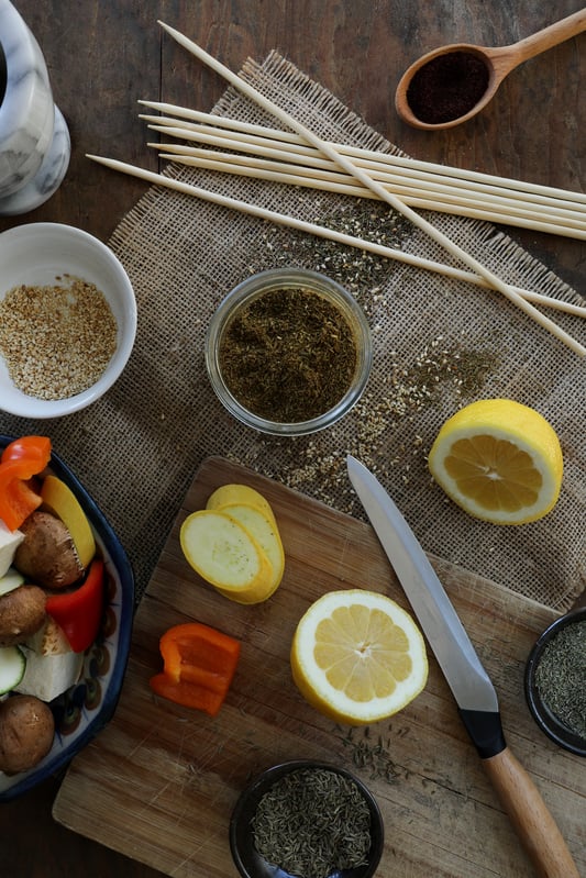 Fresh ingredients and spices are laid out on a cutting board and mat. Colorful vegetables are ready for skewering.