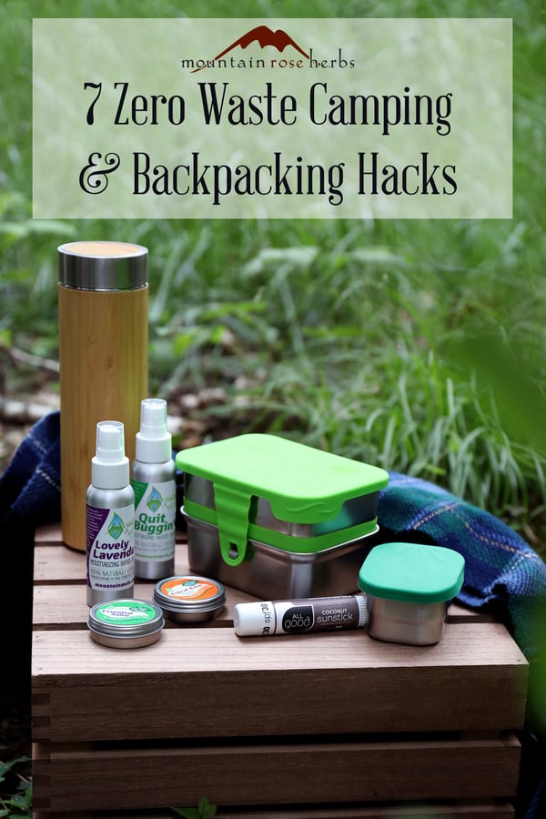 Assorted low waste camping supplies arranged on a wooden side table outdoors. Stainless steel food storage and natural outdoors products. Bamboo tea tumbler. Pinterest link to Mountain Rose Herbs. 