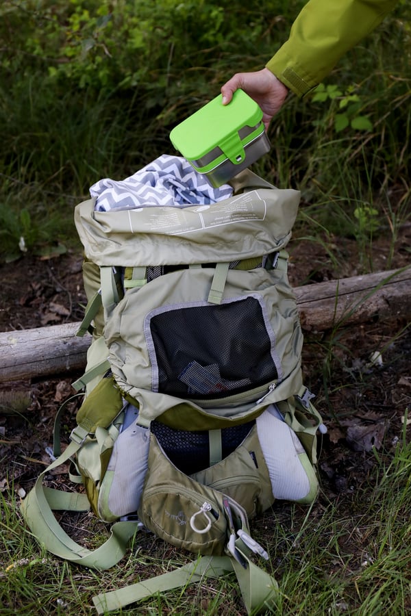 A tan hiking backpack with low waste impact being packed on a forest trail. Portable food containers are packed with first aid. 