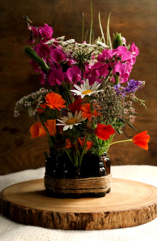 Empty amber glass bottles of different sizes are re-purposed as a centerpiece displaying colorful wildflowers and a round of pine wood. Rustic twine and wildflowers make a great seasonal centerpiece. 