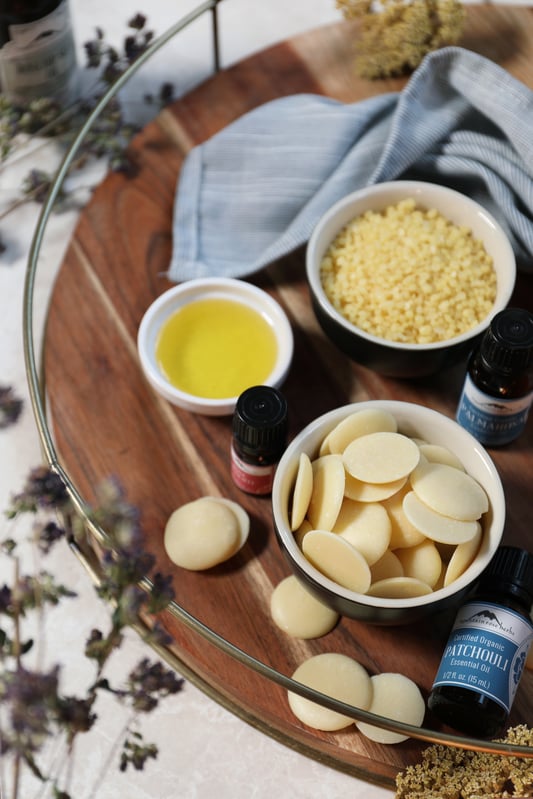 Small bowls of body care ingredients including cocoa butter wafers, oil, and beeswax pastilles.