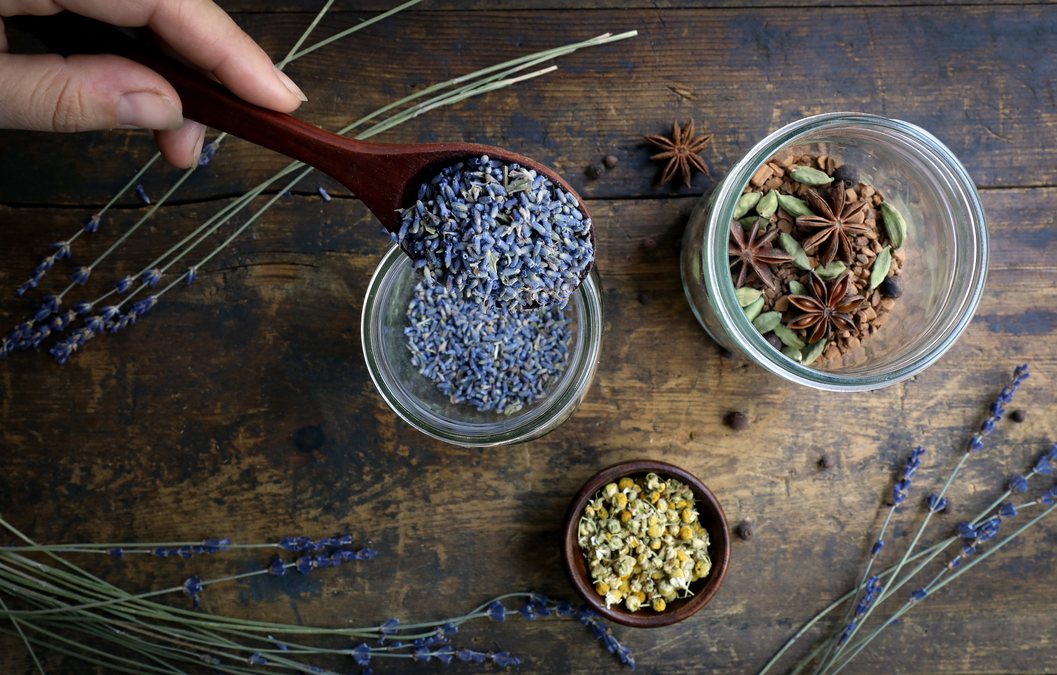 Hand pouring lavender into jar with sprigs of lavender on table and small bowl of chamomile flowers. Another jar with warming spices.