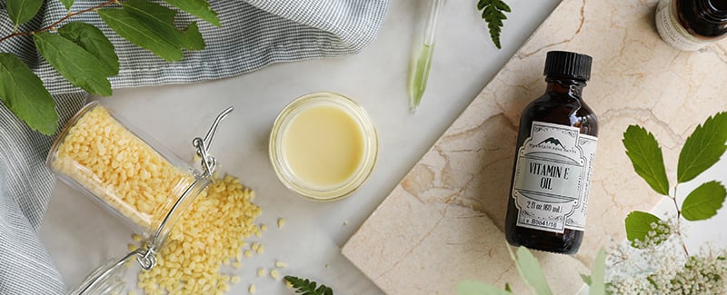 Ingredients laid out for a homemade, DIY K-Beauty skin emulsion featuring vitamin e oil, beeswax pastilles, and a finished light yellow cream.