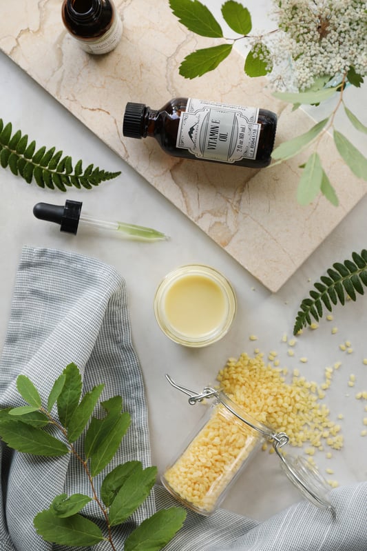 Ingredients laid out for a homemade, DIY K-Beauty skin emulsion featuring vitamin e oil, beeswax pastilles, and a finished light yellow cream.