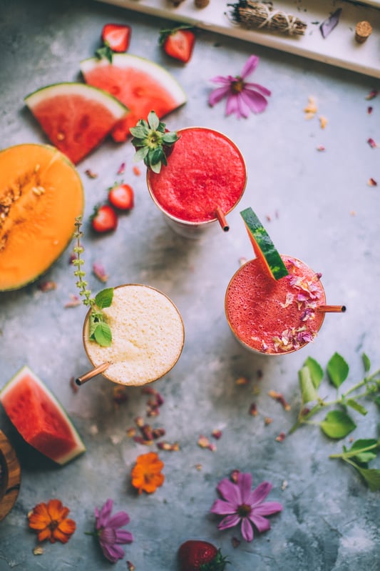 An assortment of fresh fruits, flowers, and herbs are gathered with three completed summer agua frescas garnished with strawberries, watermelon, and mint. 