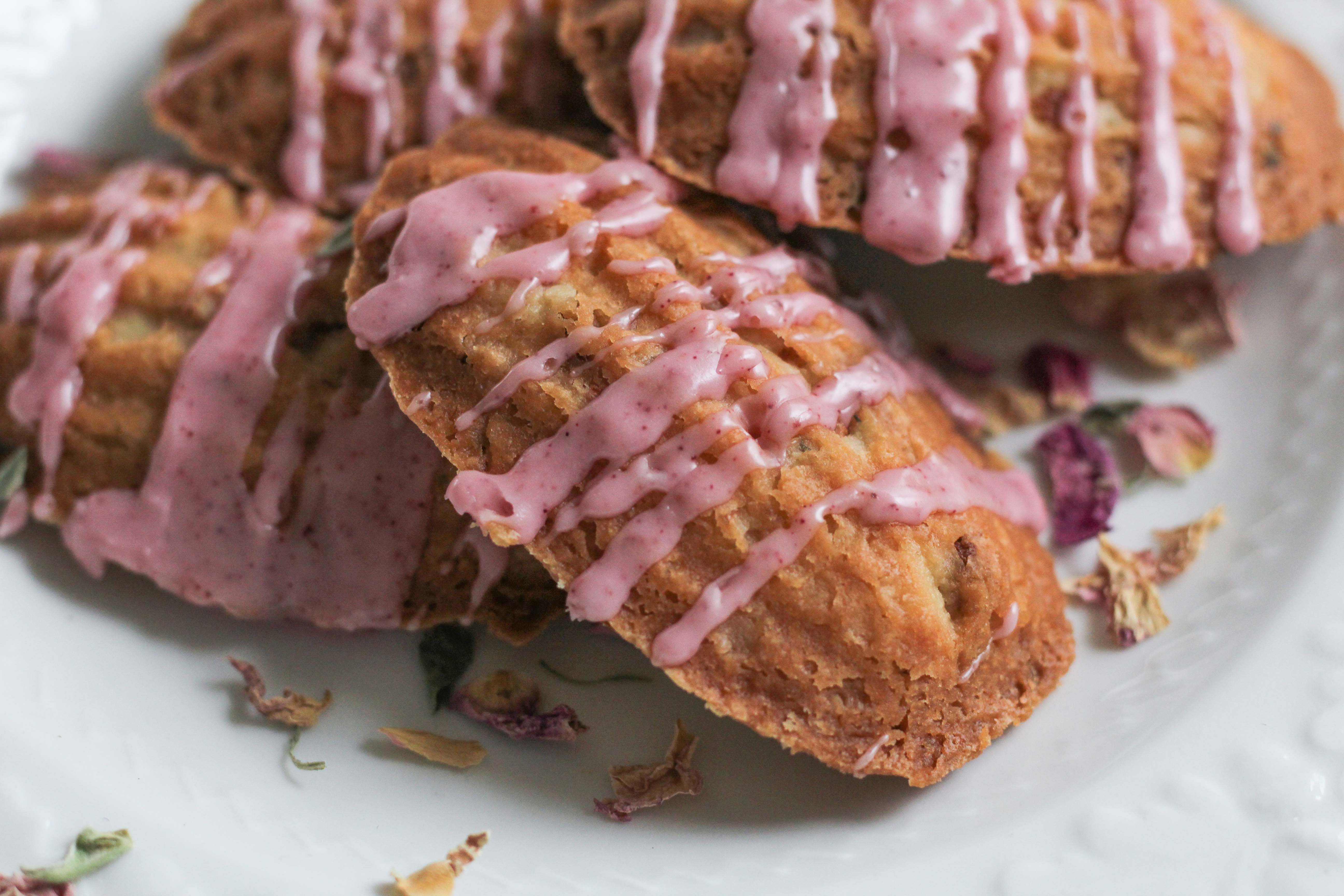 Golden Brown Madeline cookies drizzled with pink glaze displayed on white playe with rose petals displayed around it. 