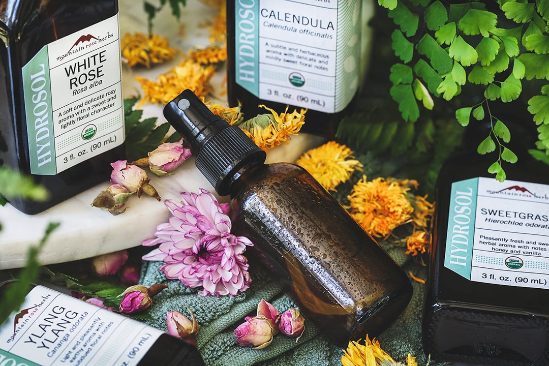 Amber glass bottle filler with DIY body toner spray surrounded by ingredients used to make it and fresh flowers.