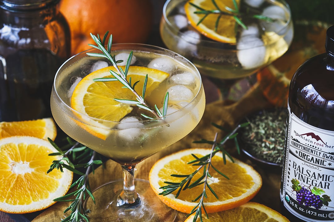 A glass of alcohol-free drink garnished with an orange slice and a sprig of rosemary surrounded by ingredients used to make it. 