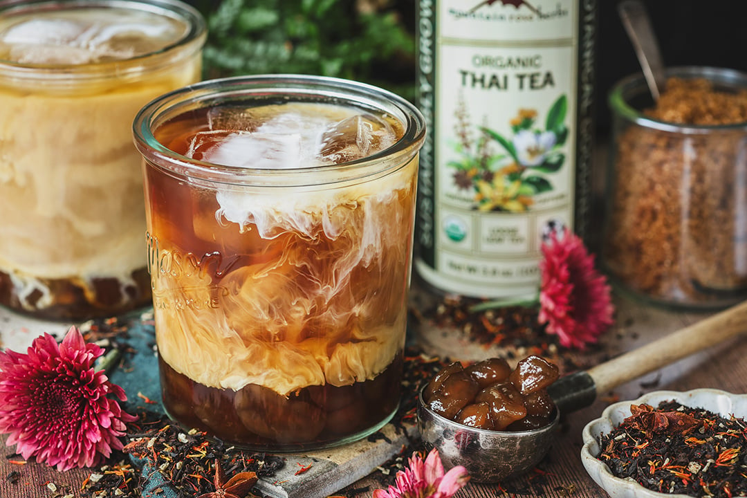 Glass with iced boba Thai tea surrounded by the tea and the tin that the tea comes in. 